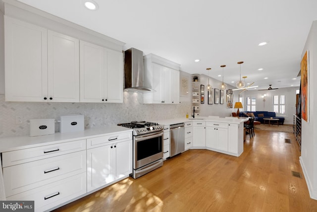kitchen featuring appliances with stainless steel finishes, white cabinets, a peninsula, open floor plan, and wall chimney exhaust hood