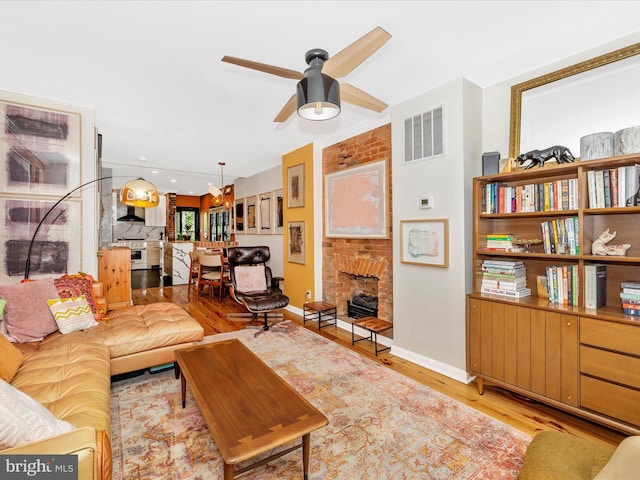 living room featuring light wood-type flooring, a ceiling fan, baseboards, and a brick fireplace