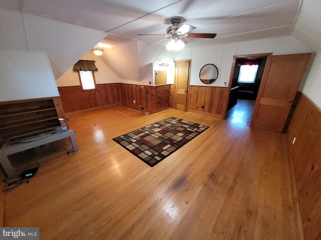 bonus room with light wood-style flooring, wooden walls, healthy amount of natural light, wainscoting, and ceiling fan