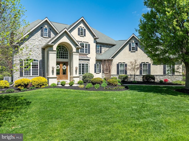 view of front facade with a front yard, stone siding, stucco siding, french doors, and central AC unit