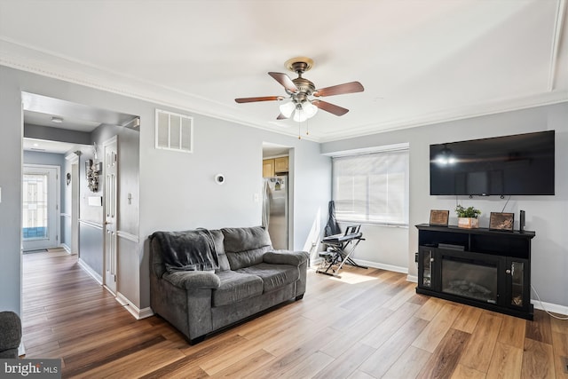living area with a ceiling fan, baseboards, light wood-type flooring, visible vents, and ornamental molding