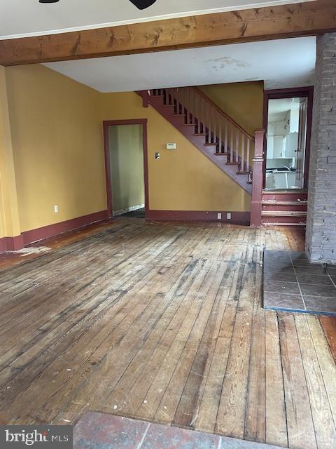 unfurnished living room with hardwood / wood-style floors, stairway, beam ceiling, and baseboards