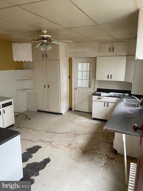 kitchen featuring white cabinetry, a sink, dark countertops, ceiling fan, and a drop ceiling