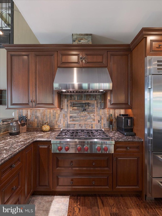 kitchen featuring dark stone countertops, under cabinet range hood, dark wood finished floors, appliances with stainless steel finishes, and backsplash