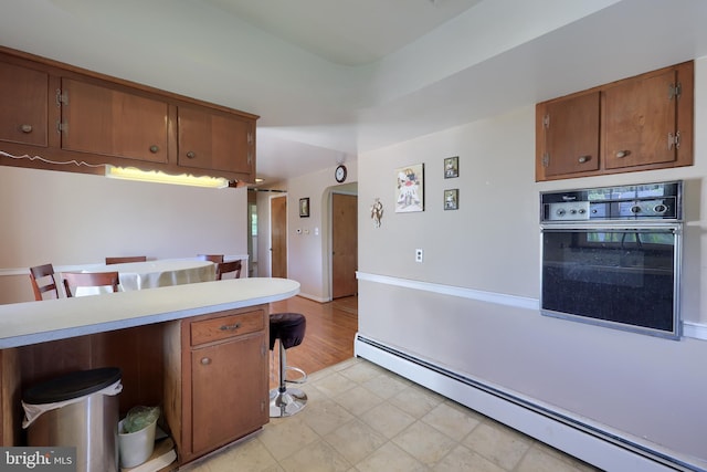 kitchen featuring brown cabinetry, baseboard heating, light countertops, a breakfast bar, and oven