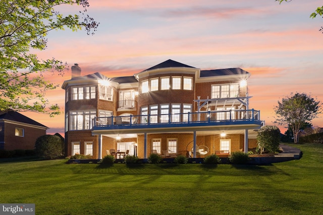 back of property at dusk featuring a lawn, ceiling fan, brick siding, and a balcony