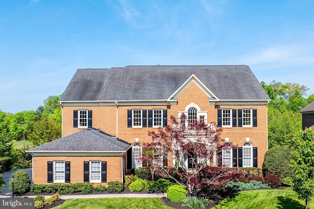 georgian-style home with roof with shingles, a front yard, and brick siding