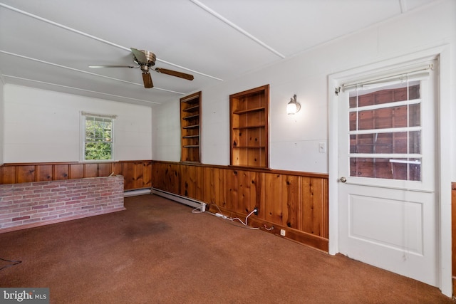 carpeted spare room with ceiling fan, wooden walls, a wainscoted wall, a baseboard radiator, and built in shelves