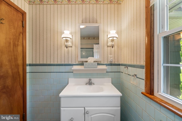 bathroom with wallpapered walls, wainscoting, vanity, and tile walls