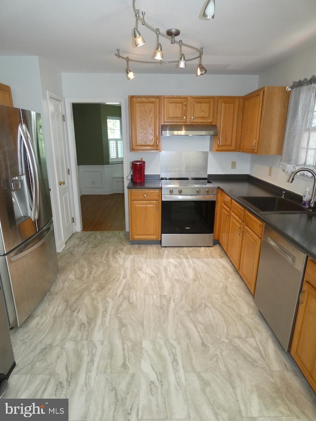 kitchen featuring brown cabinetry, dark countertops, a sink, under cabinet range hood, and stainless steel appliances