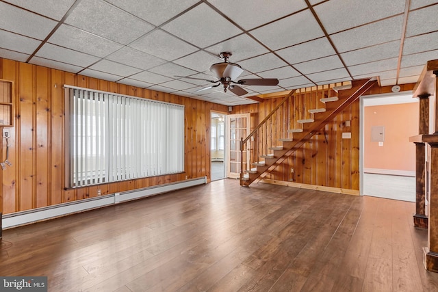 unfurnished living room featuring wood-type flooring, a ceiling fan, a baseboard heating unit, and wooden walls