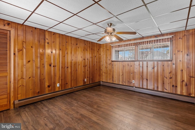 empty room featuring dark wood-style flooring, ceiling fan, a drop ceiling, and wooden walls