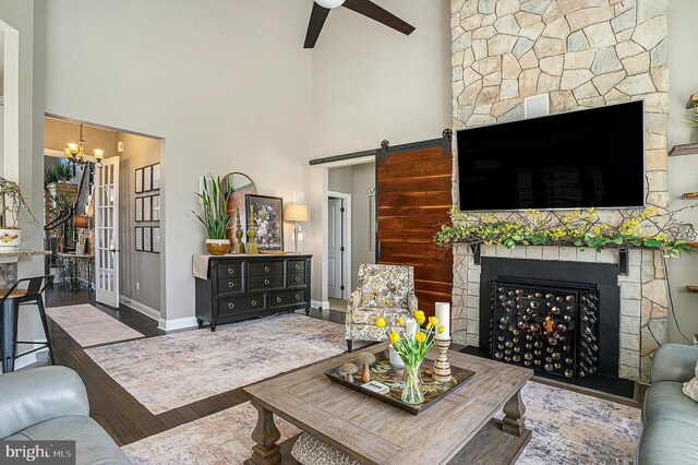 living area with a high ceiling, a barn door, a stone fireplace, ceiling fan with notable chandelier, and wood finished floors