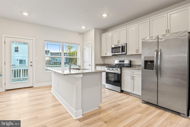 kitchen with a kitchen island with sink, light stone countertops, light wood-type flooring, a sink, and stainless steel appliances