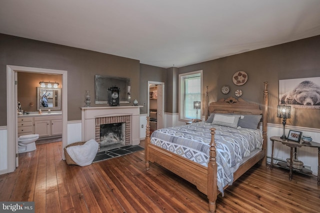 bedroom with wainscoting, hardwood / wood-style flooring, a brick fireplace, and connected bathroom