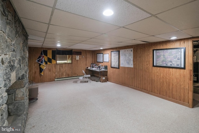 interior space with carpet flooring, wood walls, a drop ceiling, and baseboard heating