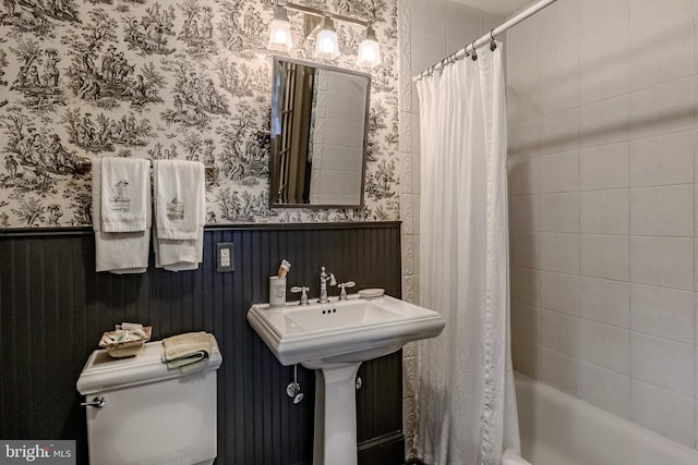 bathroom with toilet, wallpapered walls, wainscoting, and shower / tub combo with curtain