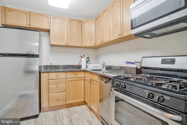 kitchen featuring appliances with stainless steel finishes, a sink, dark stone counters, light brown cabinets, and light wood-style flooring