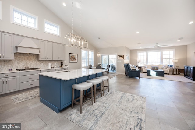 kitchen featuring a sink, high vaulted ceiling, ceiling fan with notable chandelier, custom range hood, and an island with sink