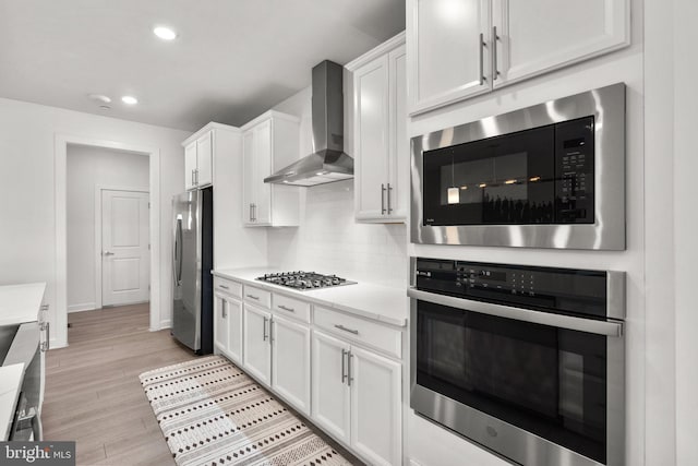 kitchen with wall chimney range hood, baseboards, light wood finished floors, stainless steel appliances, and white cabinetry