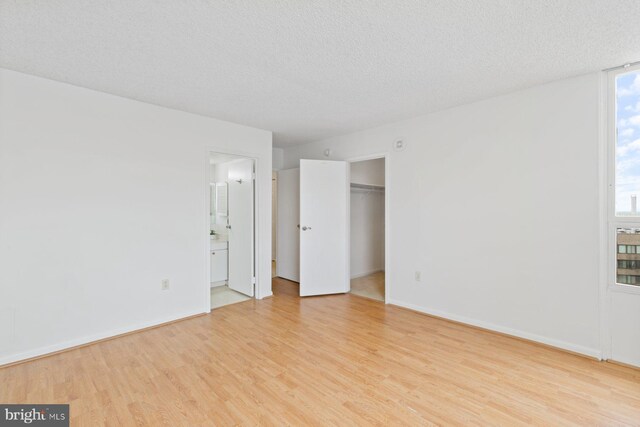 unfurnished bedroom with light wood-type flooring, a textured ceiling, baseboards, ensuite bath, and a walk in closet