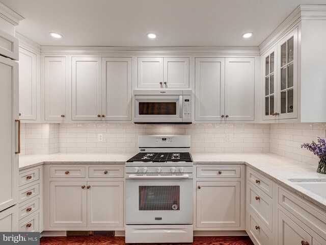 kitchen with white appliances, white cabinetry, glass insert cabinets, backsplash, and light stone countertops
