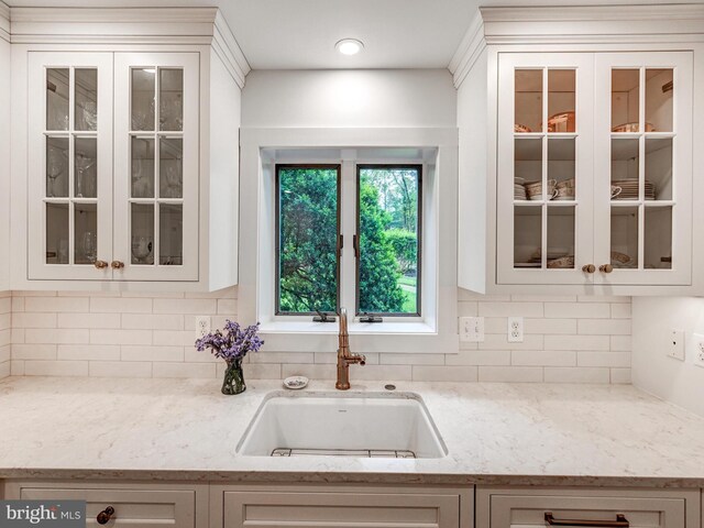 kitchen featuring a sink, backsplash, glass insert cabinets, light stone counters, and white cabinetry