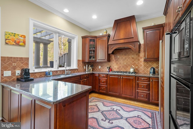 kitchen with a sink, premium range hood, black appliances, a peninsula, and dark stone counters