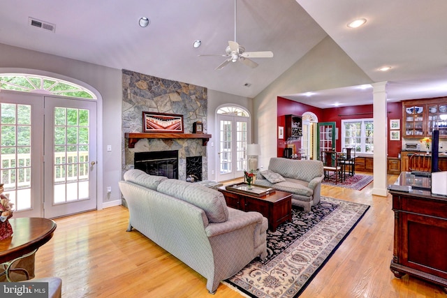 living room with ceiling fan, light wood-style floors, a stone fireplace, ornate columns, and visible vents