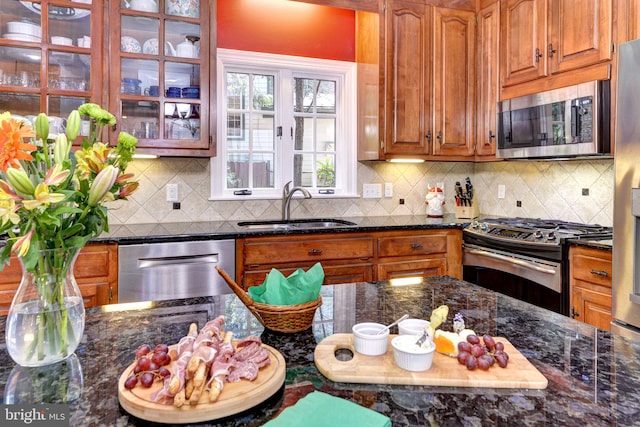 kitchen featuring appliances with stainless steel finishes, a sink, dark stone counters, tasteful backsplash, and glass insert cabinets