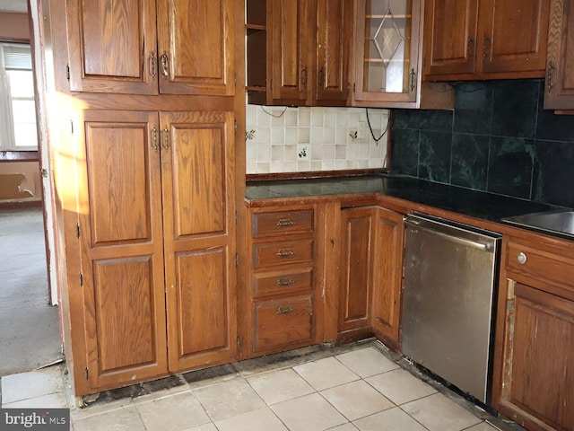 kitchen with dark countertops, brown cabinetry, decorative backsplash, stainless steel dishwasher, and glass insert cabinets