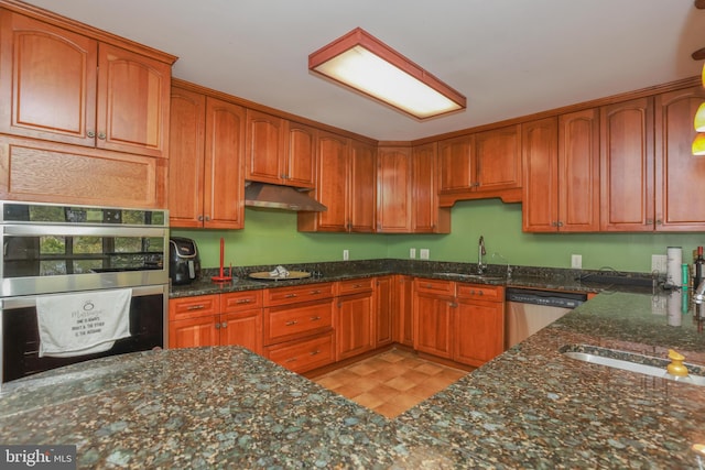 kitchen with appliances with stainless steel finishes, brown cabinets, a sink, dark stone countertops, and under cabinet range hood