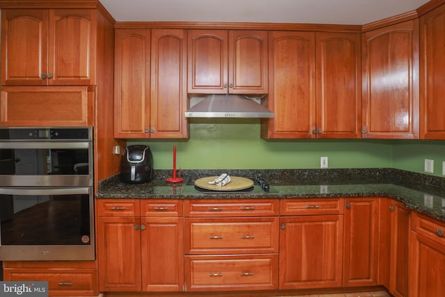 kitchen with under cabinet range hood, dark stone countertops, brown cabinetry, and double oven