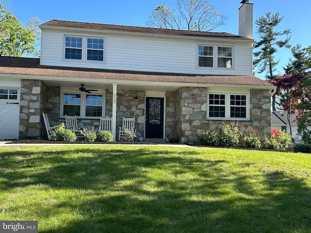 traditional home featuring stone siding, a front yard, a chimney, a ceiling fan, and covered porch