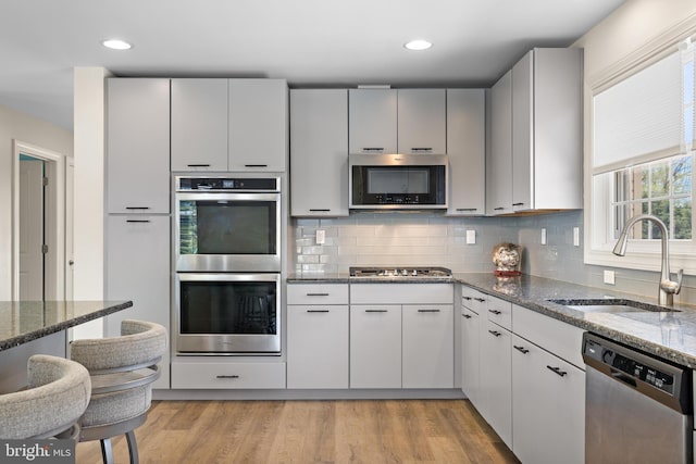 kitchen featuring light wood-style floors, tasteful backsplash, a sink, dark stone countertops, and appliances with stainless steel finishes
