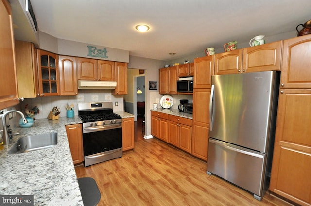 kitchen featuring glass insert cabinets, light wood-style floors, under cabinet range hood, a sink, and appliances with stainless steel finishes
