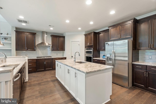 kitchen featuring appliances with stainless steel finishes, a sink, wall chimney range hood, a center island with sink, and tasteful backsplash