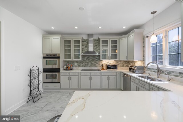 kitchen featuring wall chimney range hood, stainless steel appliances, a sink, backsplash, and light stone counters