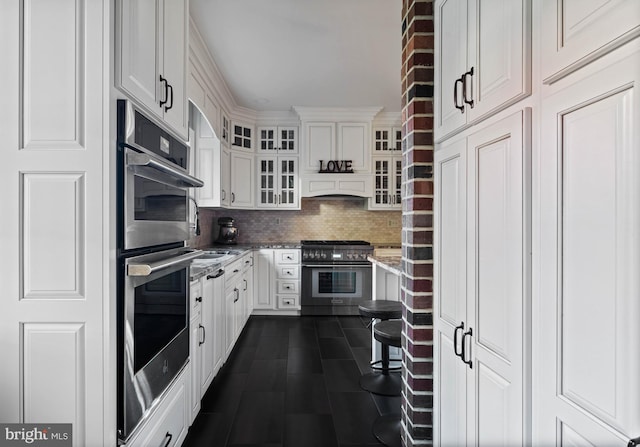 kitchen featuring white cabinets, light stone countertops, appliances with stainless steel finishes, backsplash, and glass insert cabinets