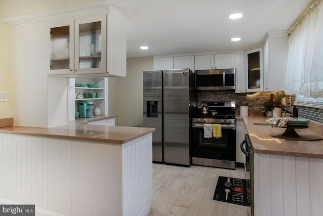 kitchen featuring appliances with stainless steel finishes, a peninsula, white cabinetry, tasteful backsplash, and glass insert cabinets