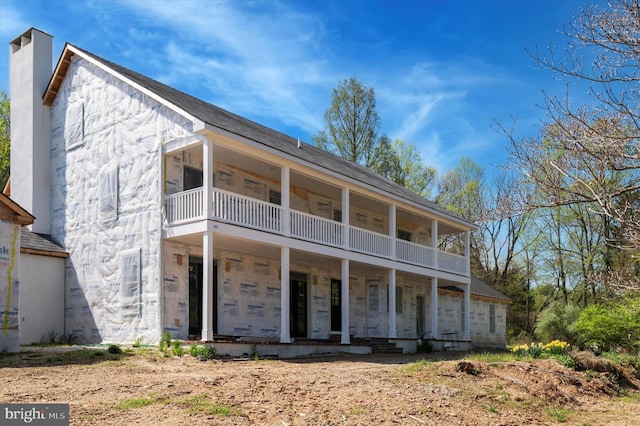 back of house with covered porch, a chimney, a balcony, and stone siding