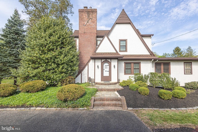 english style home with fence, a chimney, a shingled roof, and stucco siding