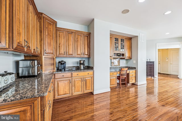 kitchen featuring brown cabinetry, wood finished floors, dark stone countertops, recessed lighting, and glass insert cabinets