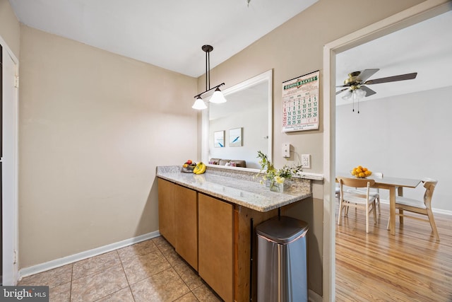 kitchen featuring brown cabinets, light stone counters, a ceiling fan, baseboards, and pendant lighting