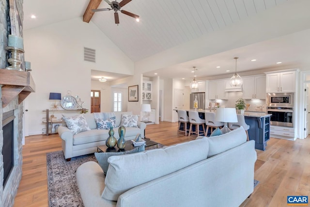 living room featuring light wood finished floors, high vaulted ceiling, a stone fireplace, a ceiling fan, and beamed ceiling
