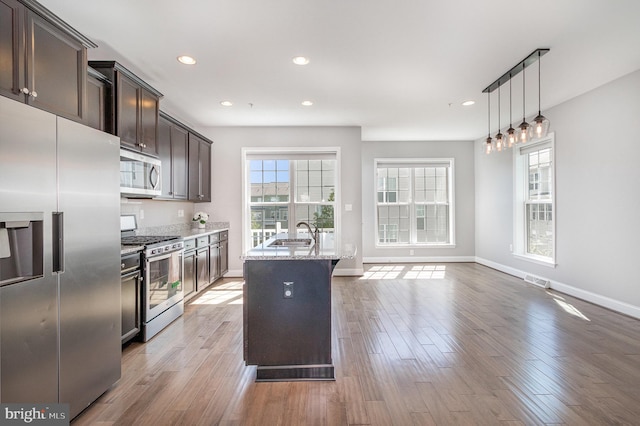 kitchen featuring dark brown cabinets, a center island with sink, light stone counters, a sink, and appliances with stainless steel finishes