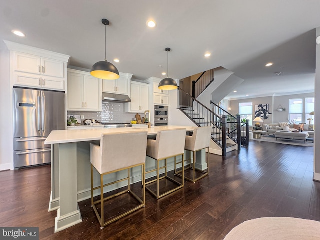 kitchen with light countertops, under cabinet range hood, a large island with sink, dark wood finished floors, and stainless steel appliances