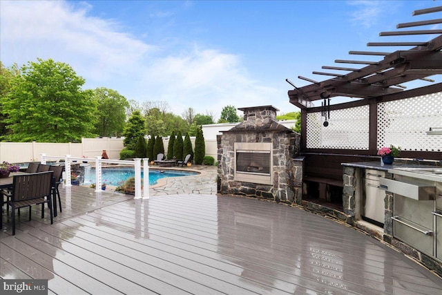 view of pool featuring an outdoor stone fireplace, a wooden deck, fence, exterior kitchen, and a pergola