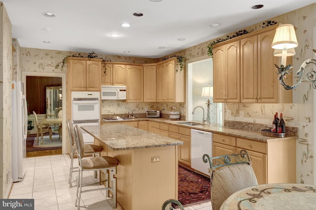 kitchen with a sink, light brown cabinets, white appliances, wallpapered walls, and a kitchen island