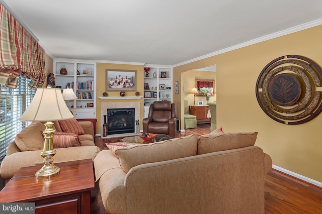 living area with ornamental molding, a glass covered fireplace, wood finished floors, baseboards, and built in shelves